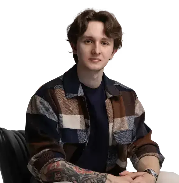 Portrait of a young man with wavy brown hair wearing a plaid overshirt over a dark t-shirt, seated with arms resting forward and tattoos visible on his forearms